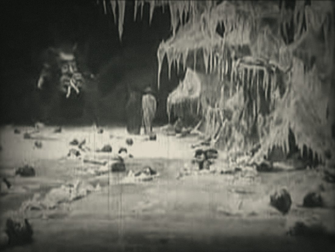 Frame from L'Inferno (1911): a cavern of Hell with bodies strewn across the ground and a demonic face watching from the dark